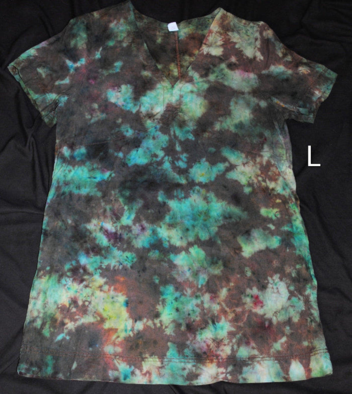 A short-sleeve tie-dye dress laid flat on a black fabric background, featuring a mottled pattern of teal, green, brown, and black. A white letter 'L' is shown to the right of the shirt.