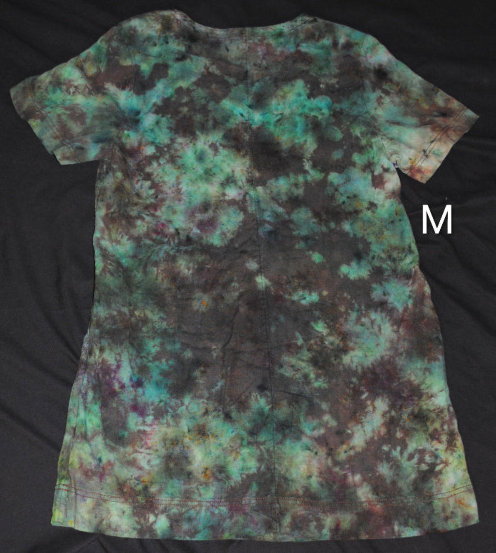 A short-sleeve tie-dye dress laid flat on a black fabric background, featuring a mottled pattern of teal, green, brown, and black. A white letter 'M' is shown to the right of the shirt.