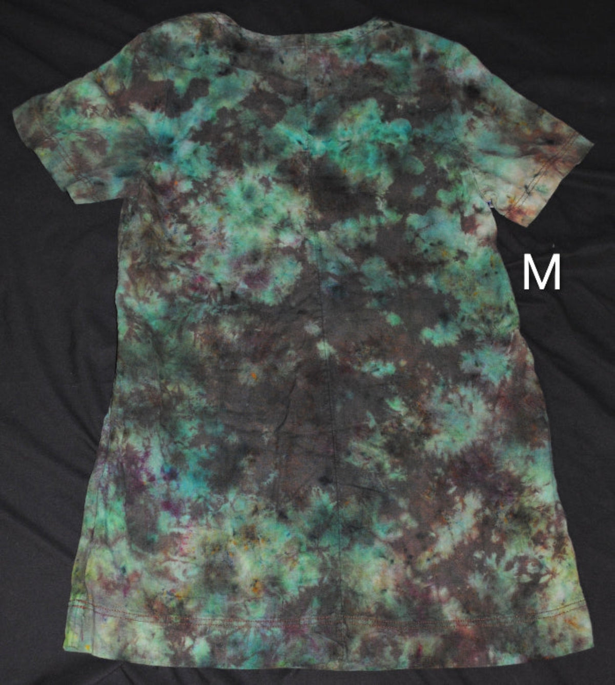 A short-sleeve tie-dye dress laid flat on a black fabric background, featuring a mottled pattern of teal, green, brown, and black. A white letter 'M' is shown to the right of the shirt.