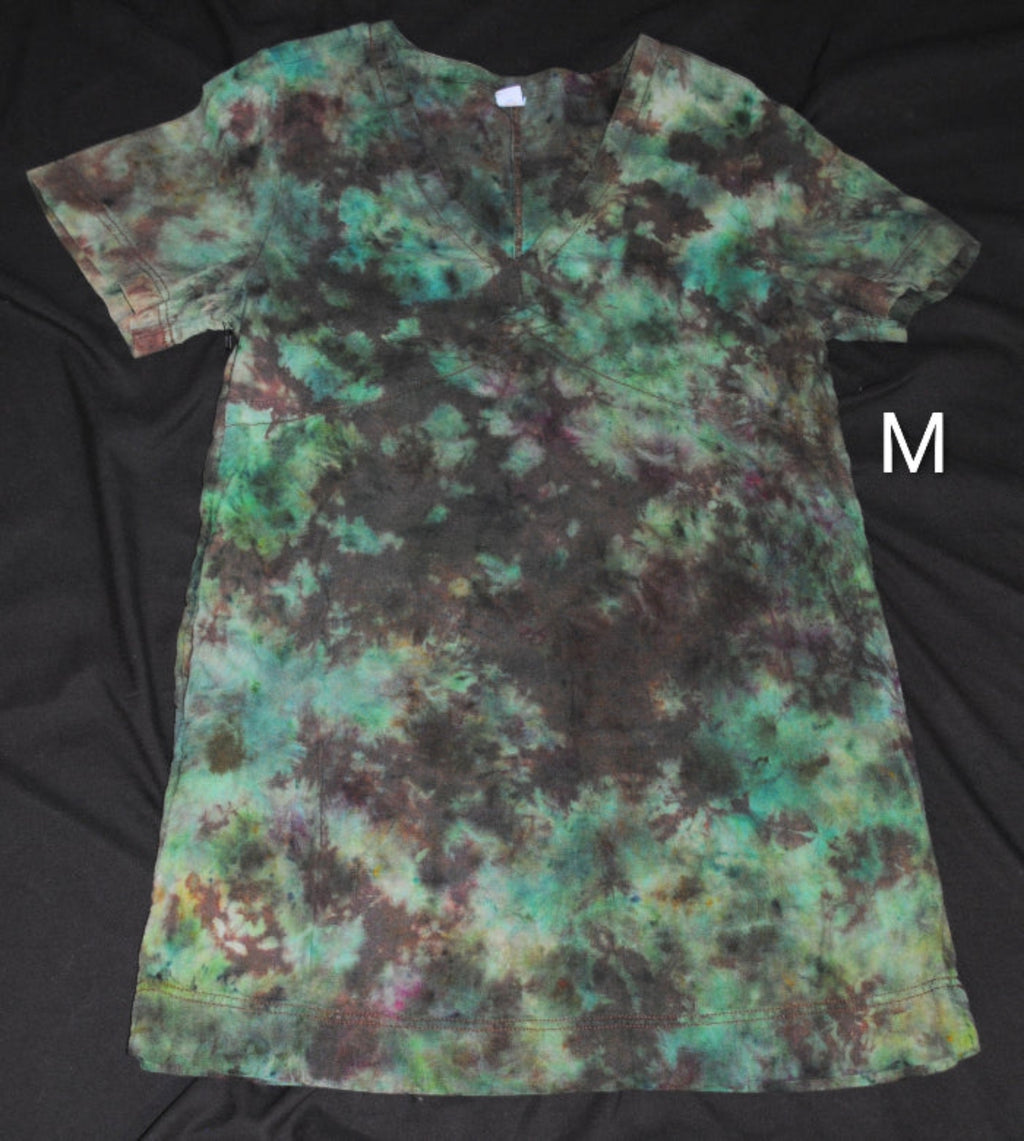 A short-sleeve tie-dye dress laid flat on a black fabric background, featuring a mottled pattern of teal, green, brown, and black. A white letter 'M' is shown to the right of the shirt.