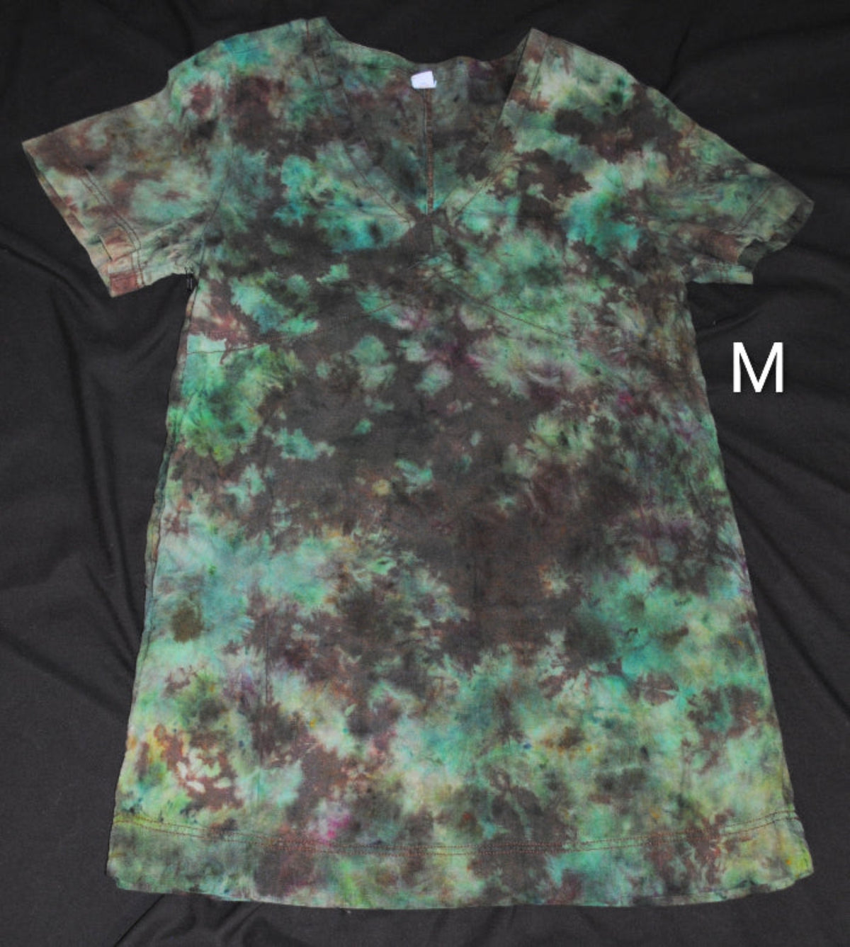 A short-sleeve tie-dye dress laid flat on a black fabric background, featuring a mottled pattern of teal, green, brown, and black. A white letter 'M' is shown to the right of the shirt.