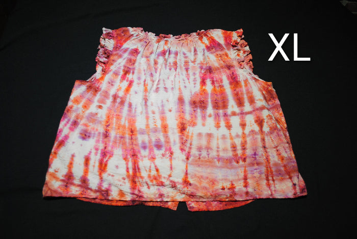 A sleeveless tie-dye top laid flat on a black background, featuring a white base with orange, pink, and purple vertical tie-dye streaks. The word 'XL' is printed in white on the upper right.