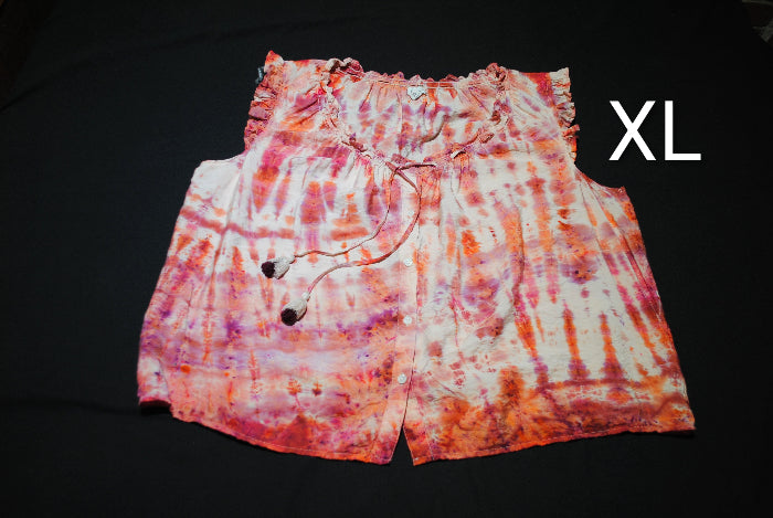 A sleeveless white tank top with a pink and orange tie-dye pattern, featuring a ruffled neckline, drawstring waist, and snap buttons, laid flat on a black background with an 'XL' size label overlay.