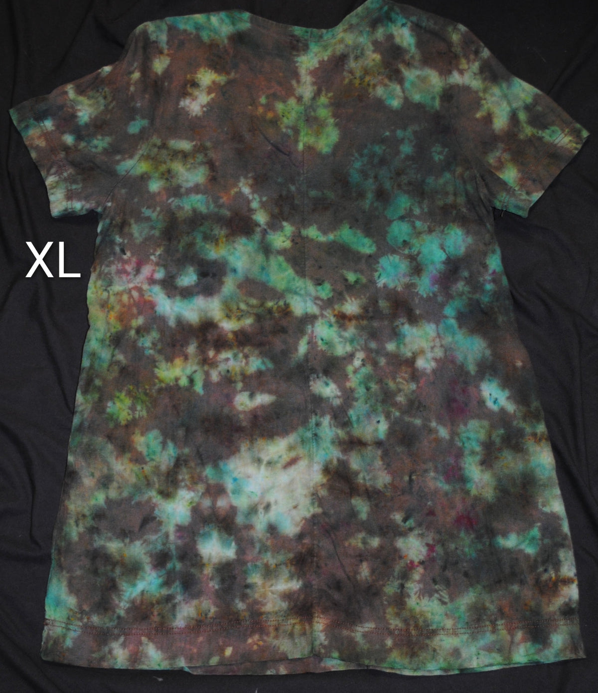 A short-sleeve tie-dye dress laid flat on a black fabric background, featuring a mottled pattern of teal, green, brown, and black. White letters 'XL' is shown to the left of the dress.