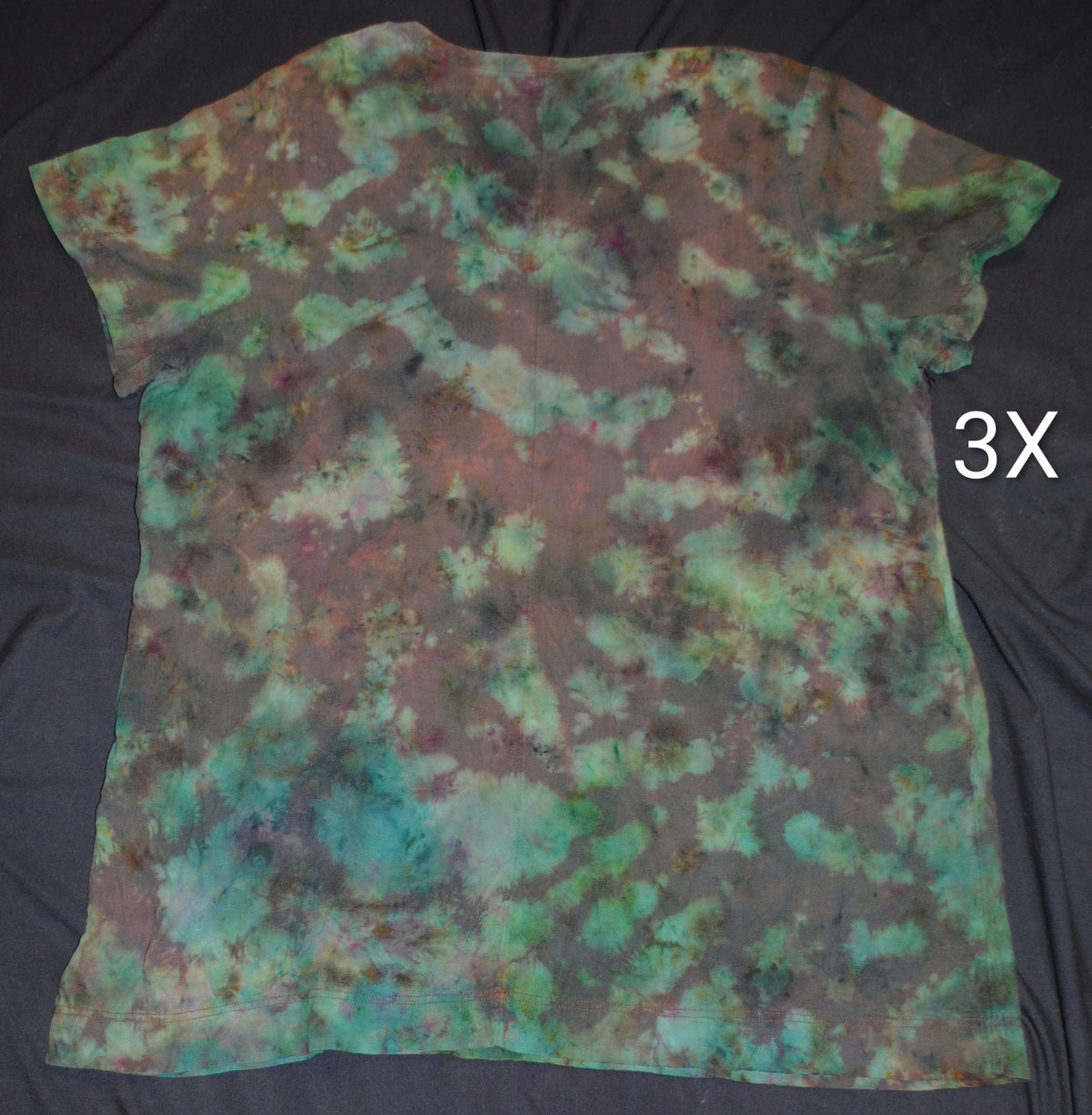 A short-sleeve tie-dye dress laid flat on a black fabric background, featuring a mottled pattern of teal, green, brown, and black. White letters '3X' is shown to the right of the shirt.