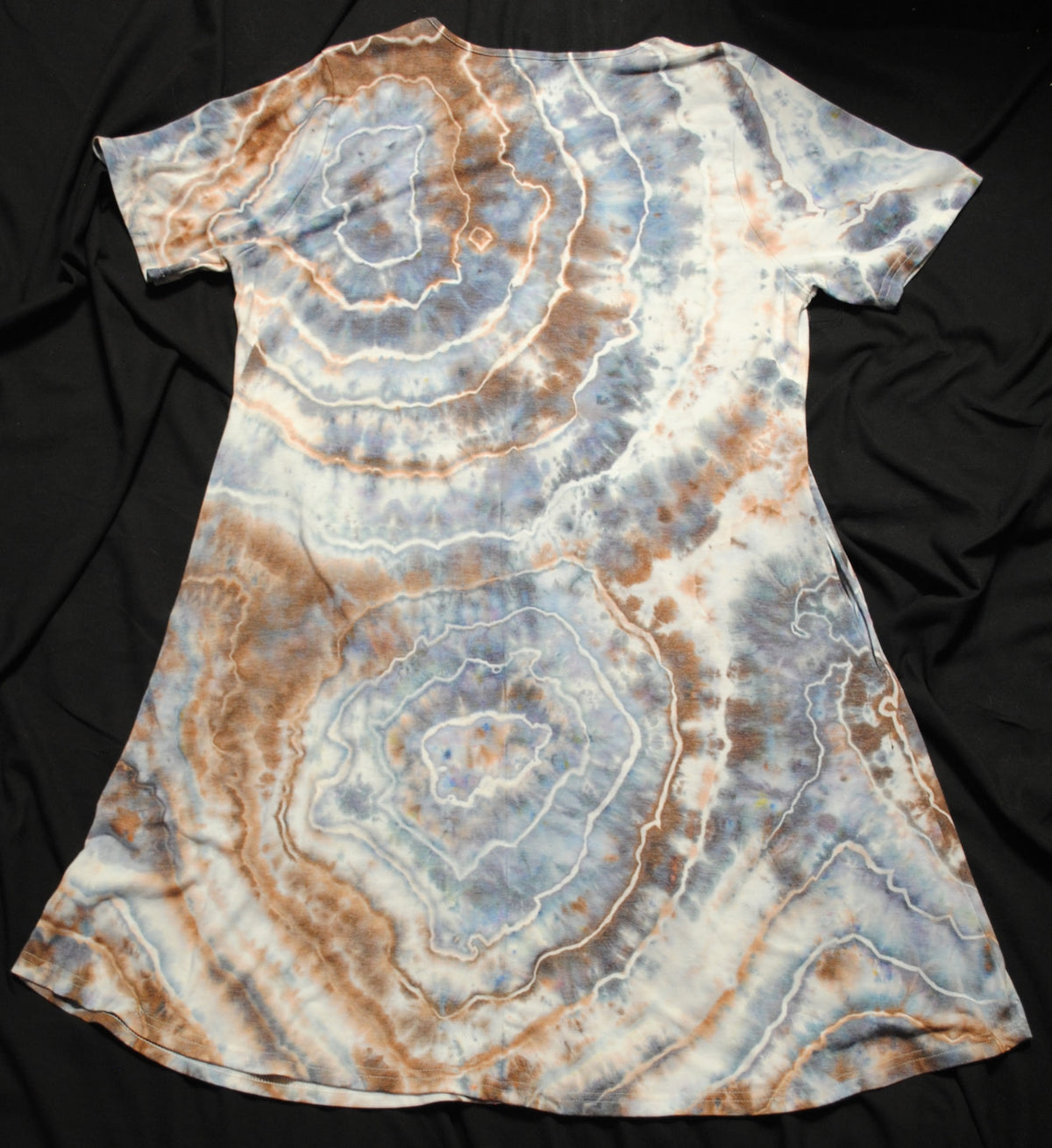 A short-sleeve tie-dye tunic dress with concentric circle patterns in light blue, brown, and white, laid flat on a black fabric background.