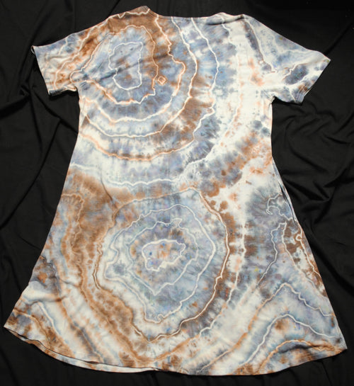 A short-sleeve tie-dye tunic dress with concentric circle patterns in light blue, brown, and white, laid flat on a black fabric background.