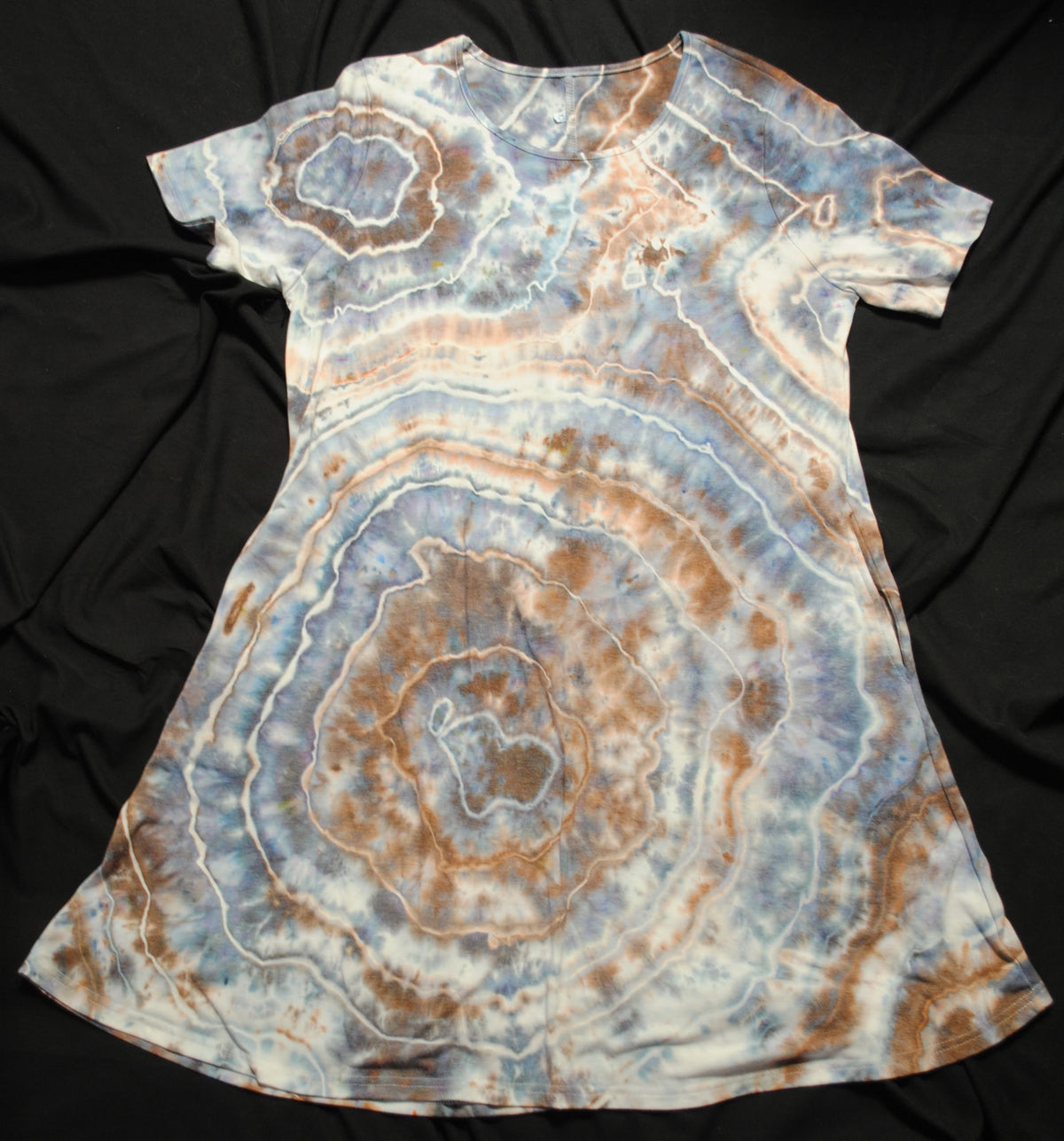 A short-sleeve tie-dye dress laid flat on a black fabric background. The dress features a concentric agate or geode-like pattern in shades of light blue, white, and warm brown.