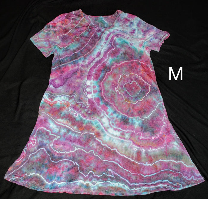 A short-sleeve tie-dye dress laid flat on a black background, featuring a spiral pattern in pink, purple, teal, and white. A white letter 'M' is printed on the right side.