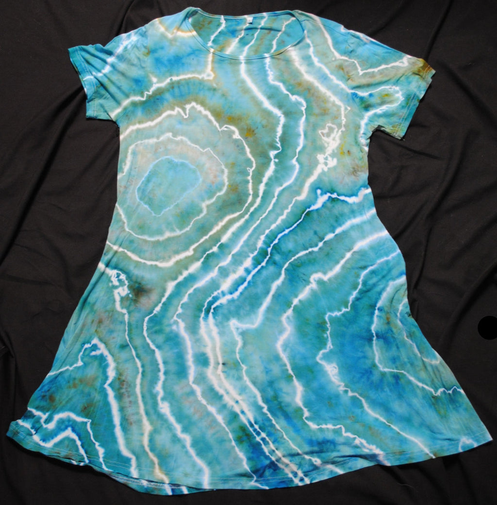 A short-sleeve tie-dye t-shirt dress with a blue, teal, and white agate-like geode pattern, laid flat on a black fabric background.